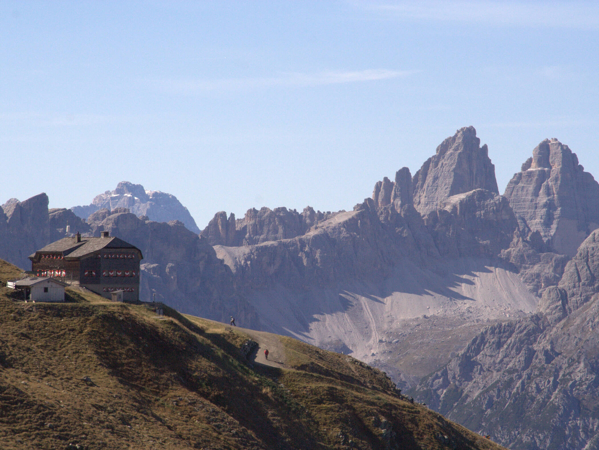 Eine Berghütte liegt auf einem grasbewachsenen Bergrücken mit den zerklüfteten Gipfeln der Dolomiten im Hintergrund. - AMIL chalets dolomites
