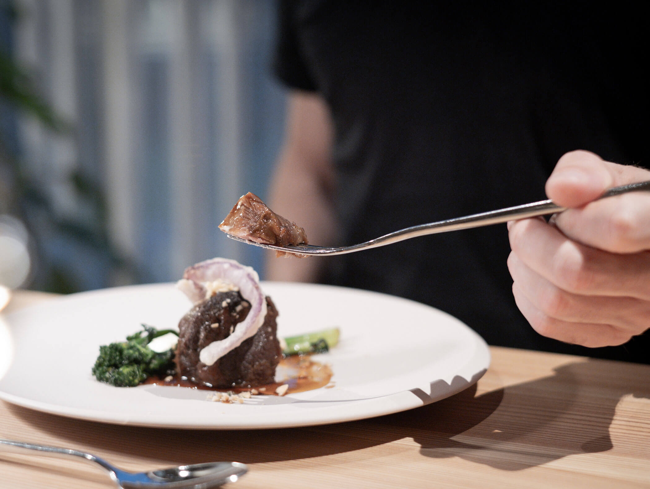Una persona che tiene in mano una forchetta con un boccone di carne sopra un piatto gourmet impiattato su un tavolo di legno. - AMIL chalets dolomites