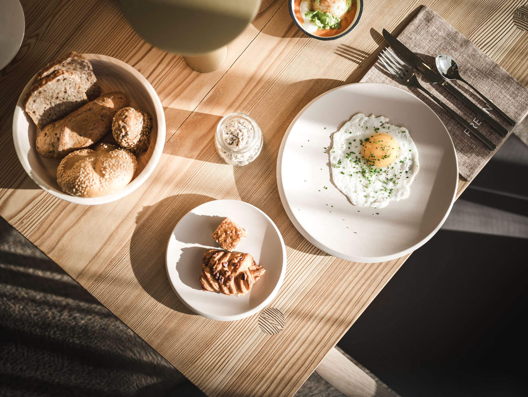 Un tavolo per la colazione con un uovo fritto, panini, pasticcini e utensili su una superficie di legno. - AMIL chalets dolomites