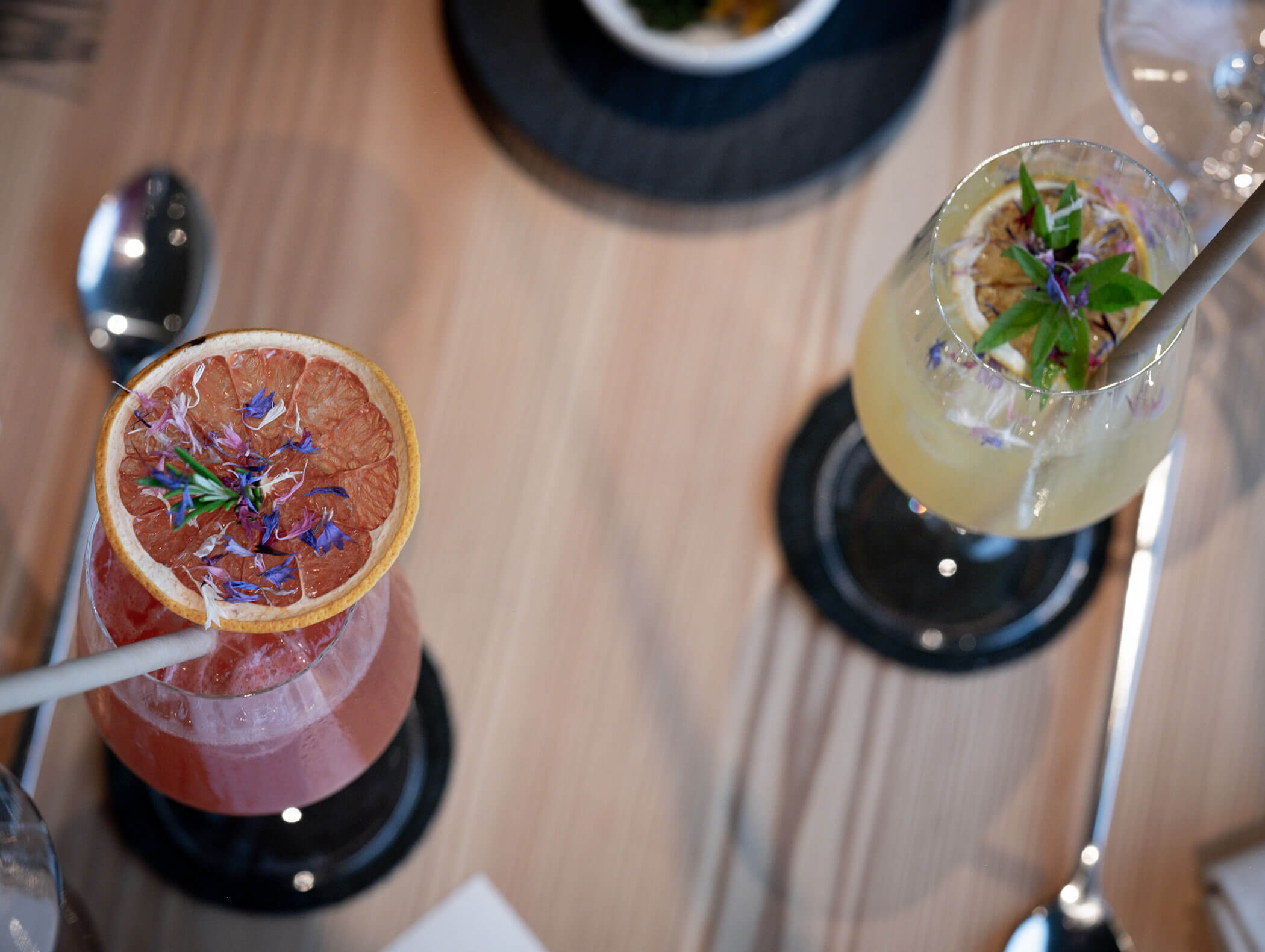 Due cocktail colorati guarniti con agrumi ed erbe essiccate su un tavolo di legno, visti dall'alto. - AMIL chalets dolomites