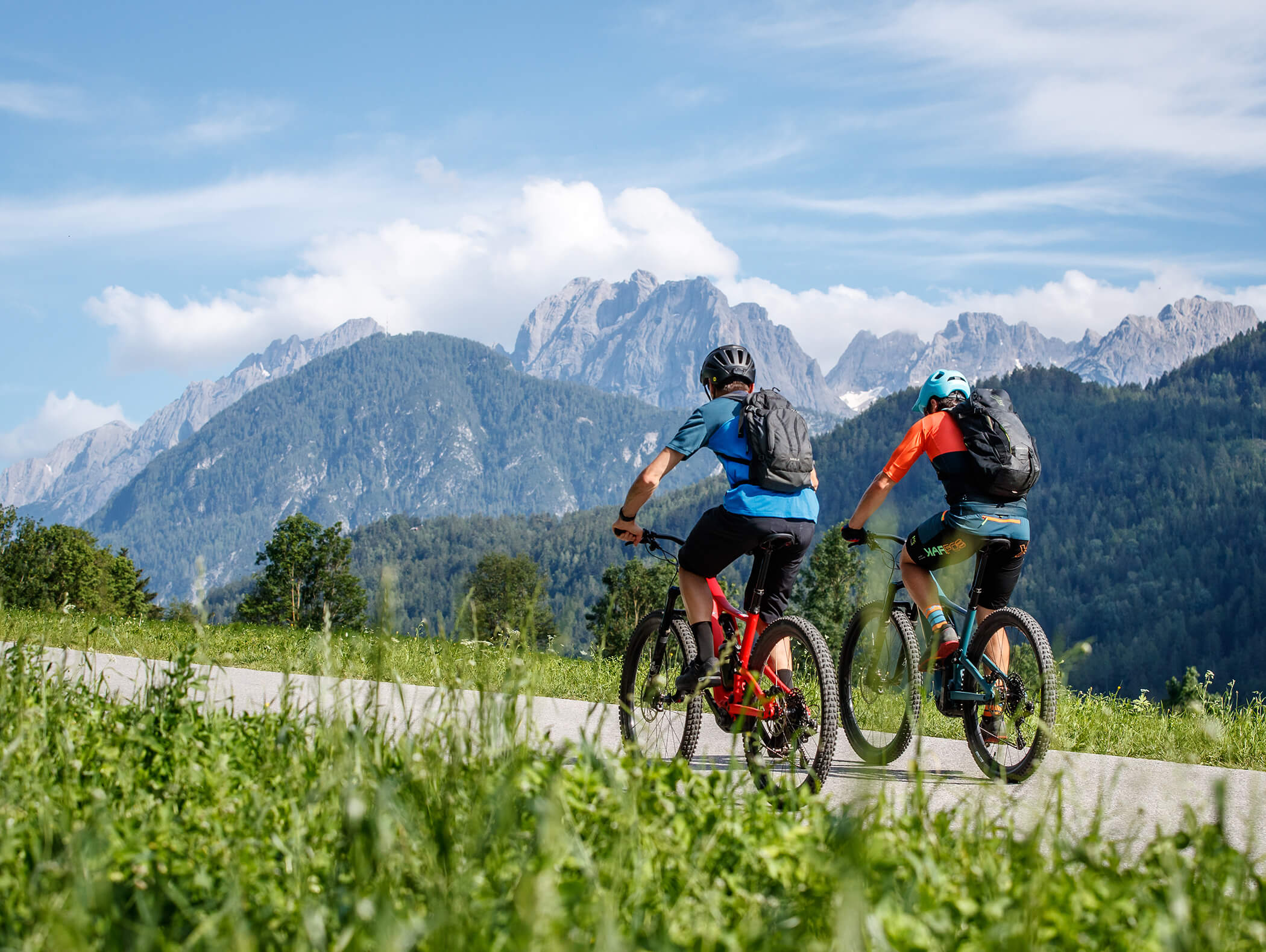 Zwei Radfahrer fahren auf einem Weg durch grüne Felder mit Bergen und blauem Himmel im Hintergrund. - AMIL chalets dolomites