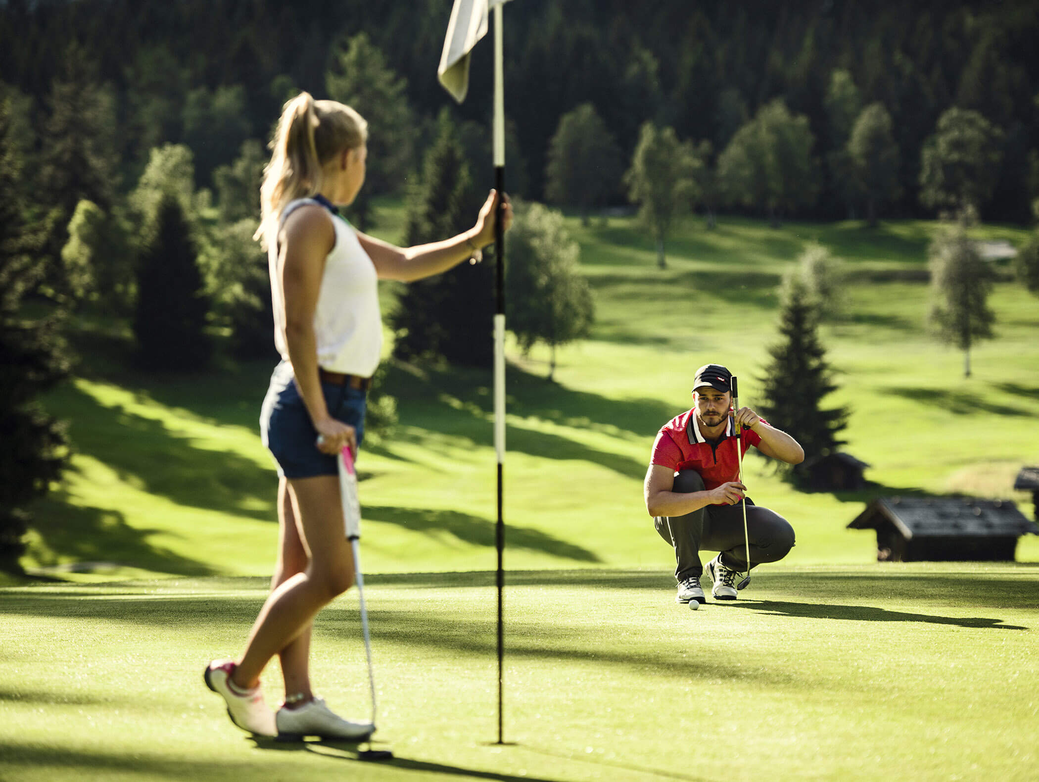 Eine Frau hält einen Flaggenstock, während ein Mann in der Hocke auf einem sonnigen Golfplatz einen Putt anlegt. - AMIL chalets dolomites