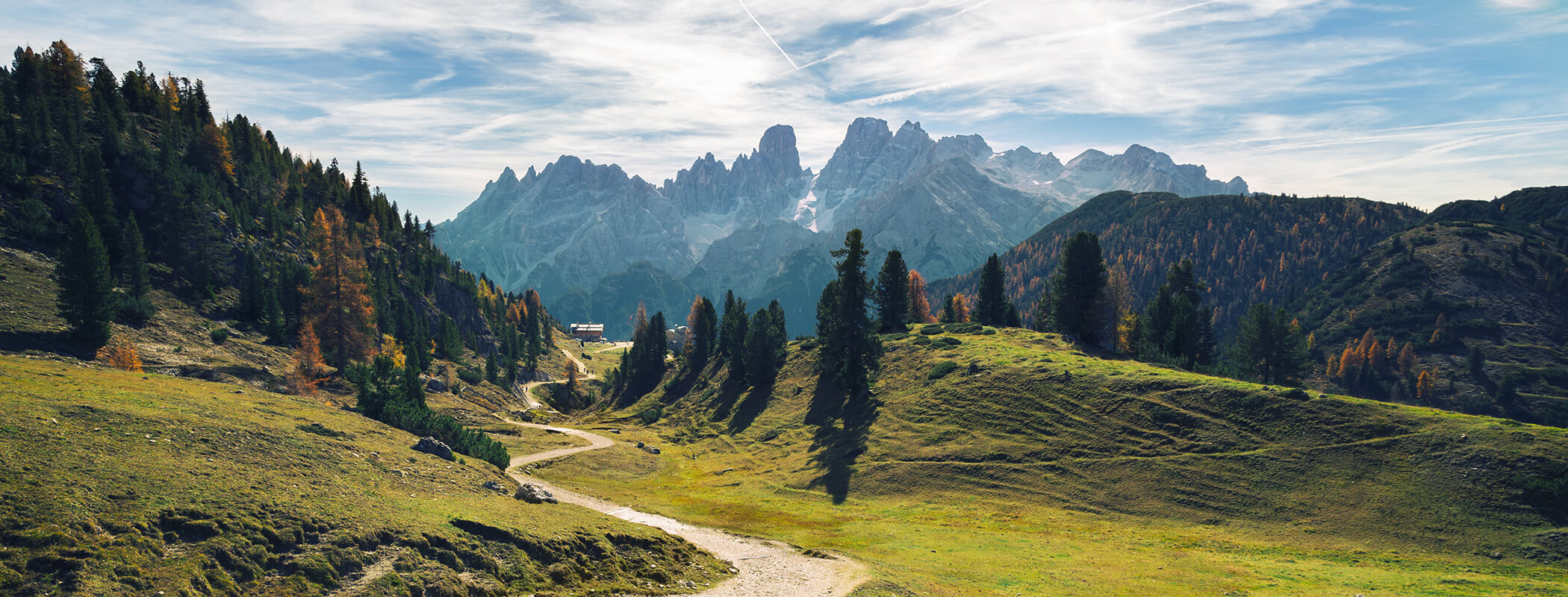 Ein gewundener Feldweg führt durch grüne Hügel zu hohen, felsigen Bergen unter einem blauen, wolkenverhangenen Himmel. - AMIL chalets dolomites