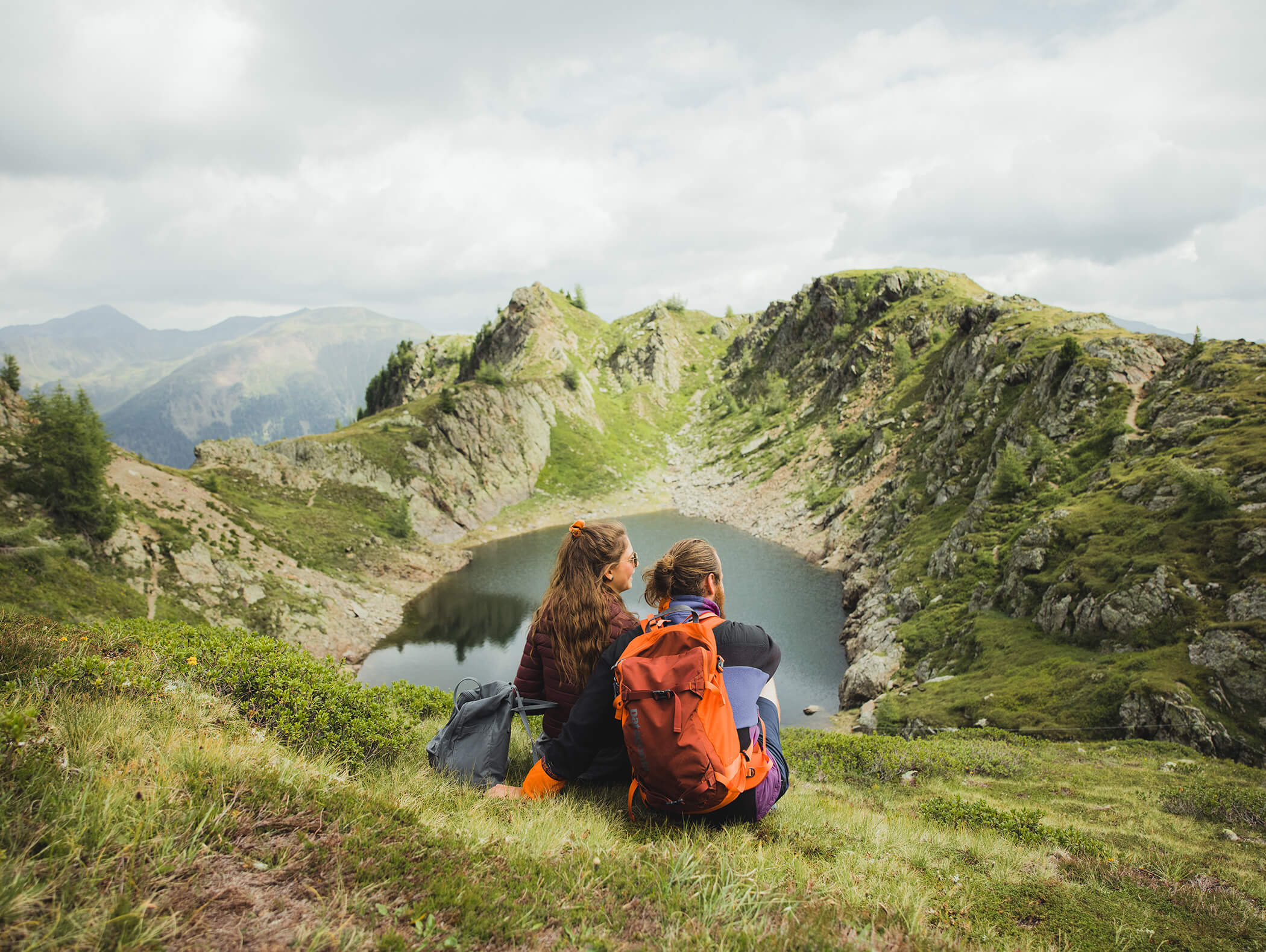 Zwei Personen mit Rucksäcken sitzen im Gras und blicken auf einen Bergsee, der von felsigen Hügeln und wolkenverhangenem Himmel umgeben ist. - AMIL chalets dolomites