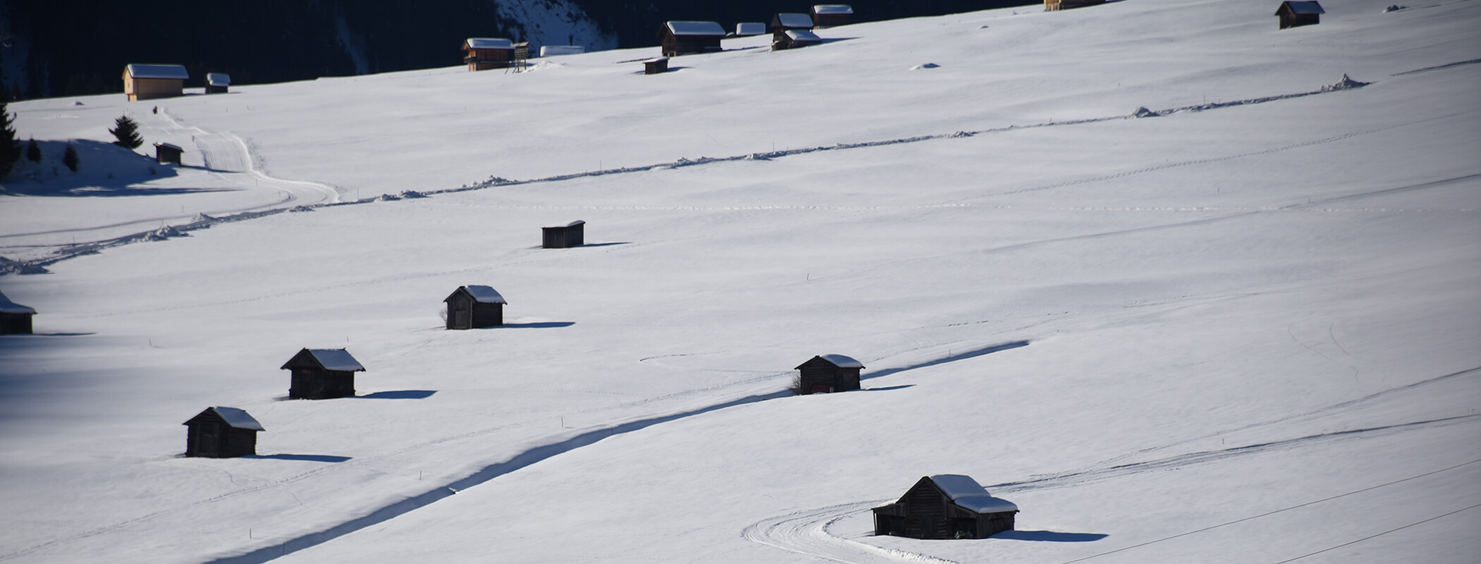 Kleine Holzhütten, verstreut über einen schneebedeckten Hügel im hellen Sonnenlicht. - AMIL chalets dolomites
