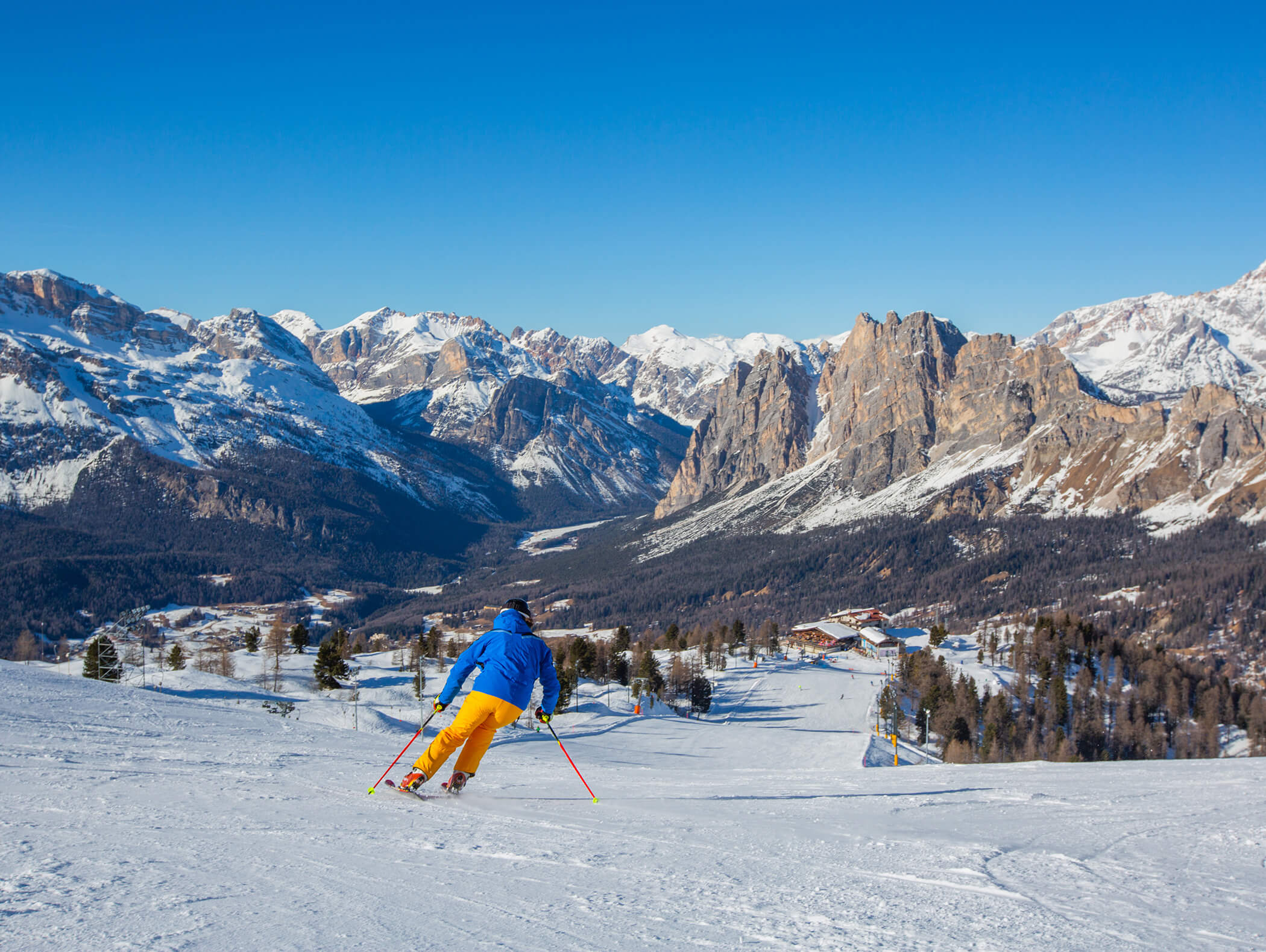 Ein Skifahrer in Blau und Gelb fährt einen verschneiten Hang hinunter, mit Bergen und klarem blauen Himmel im Hintergrund. - AMIL chalets dolomites