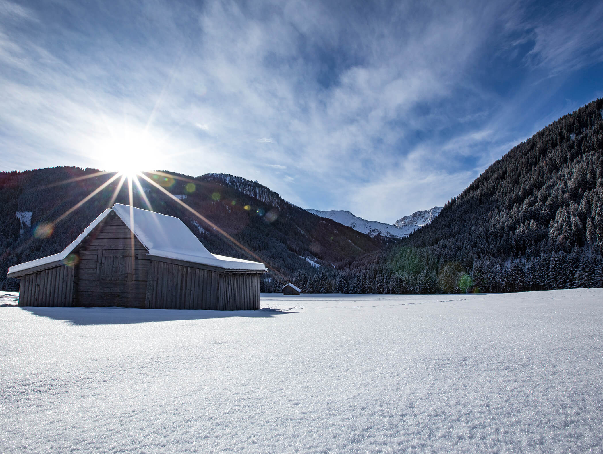 Eine schneebedeckte Hütte auf einem verschneiten Feld mit Bergen und Sonnenschein im Hintergrund. - AMIL chalets dolomites