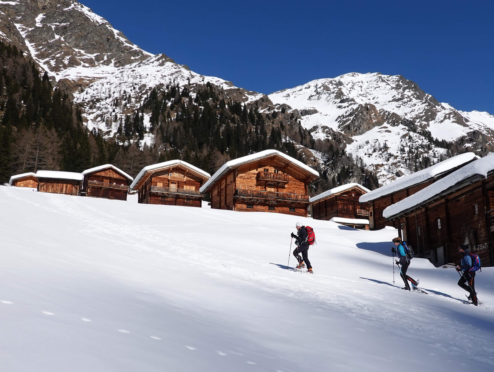 Zwei Personen mit Rucksäcken wandern einen verschneiten Hang in der Nähe von Holzhütten und schneebedeckten Bergen hinauf. - AMIL chalets dolomites