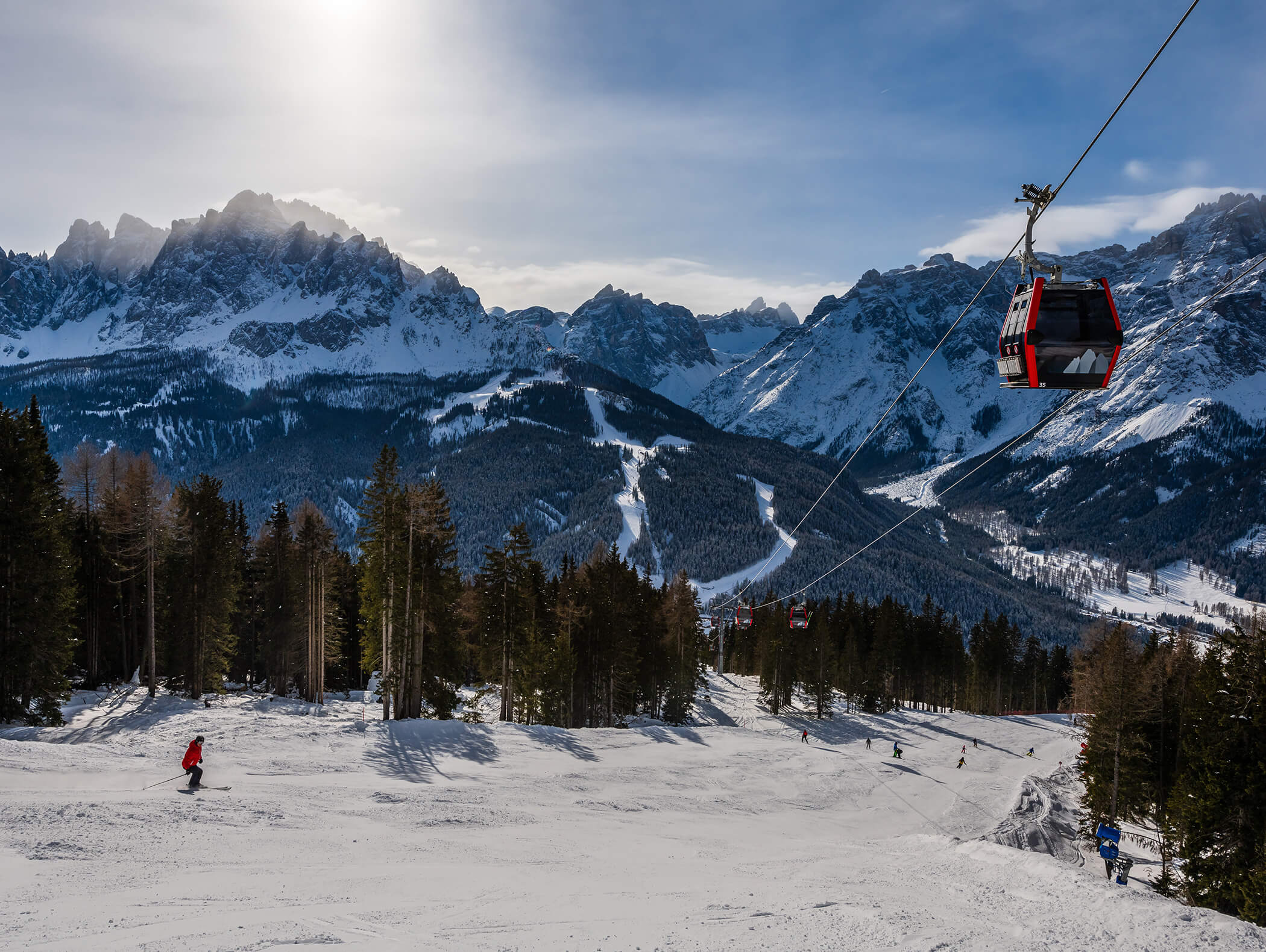 Skifahrer und eine rote Gondel auf einem verschneiten Berghang mit Wald und Gipfeln im Hintergrund. - AMIL chalets dolomites