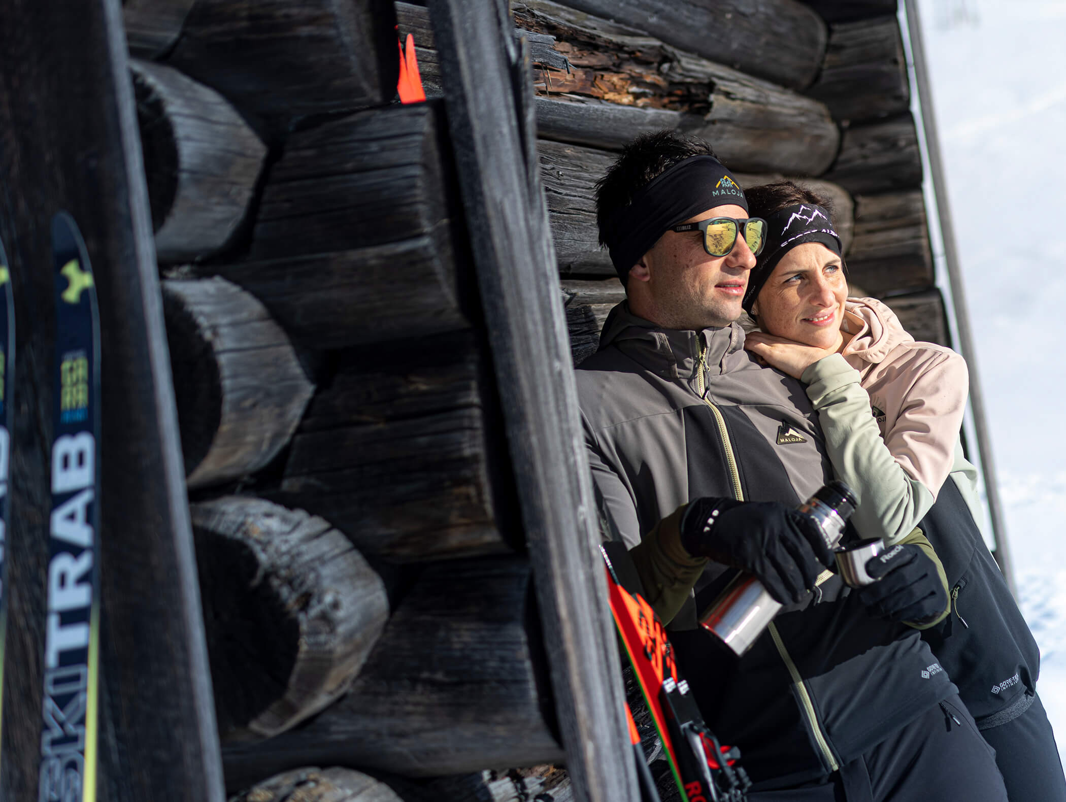 Zwei Personen in Winterkleidung stehen an einer Blockhütte und genießen die verschneite Natur mit Skiern und einer Thermoskanne. - AMIL chalets dolomites
