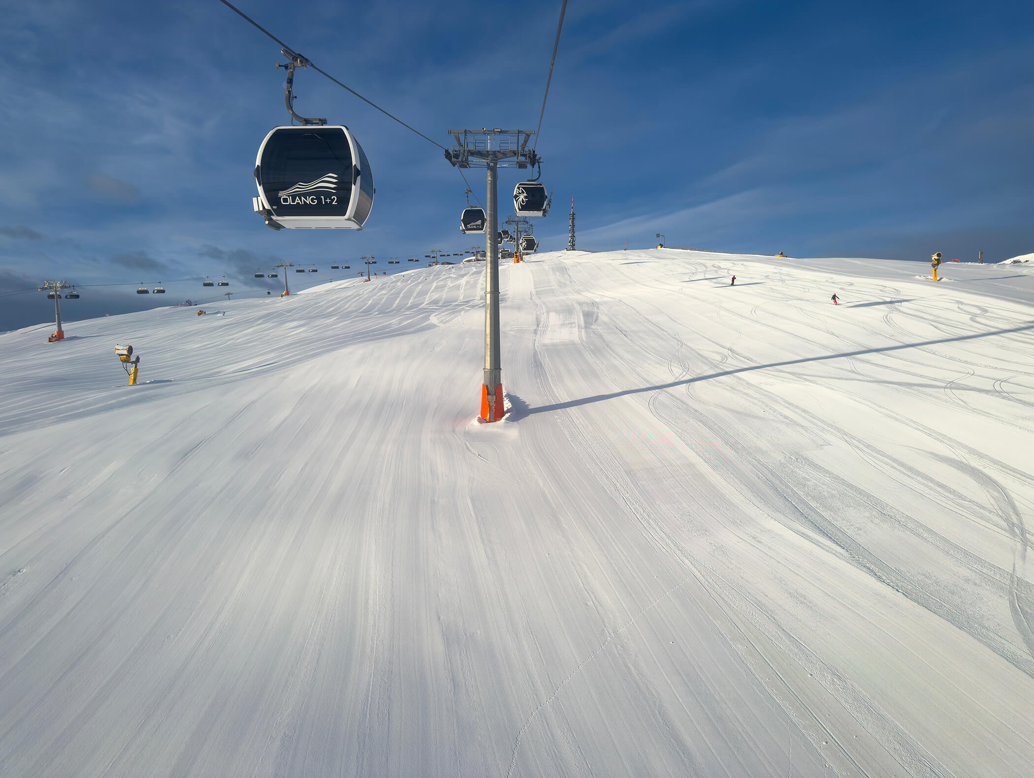 Skiliftgondeln fahren über eine verschneite Skipiste unter blauem Himmel mit ein paar Skifahrern in der Ferne. - AMIL chalets dolomites