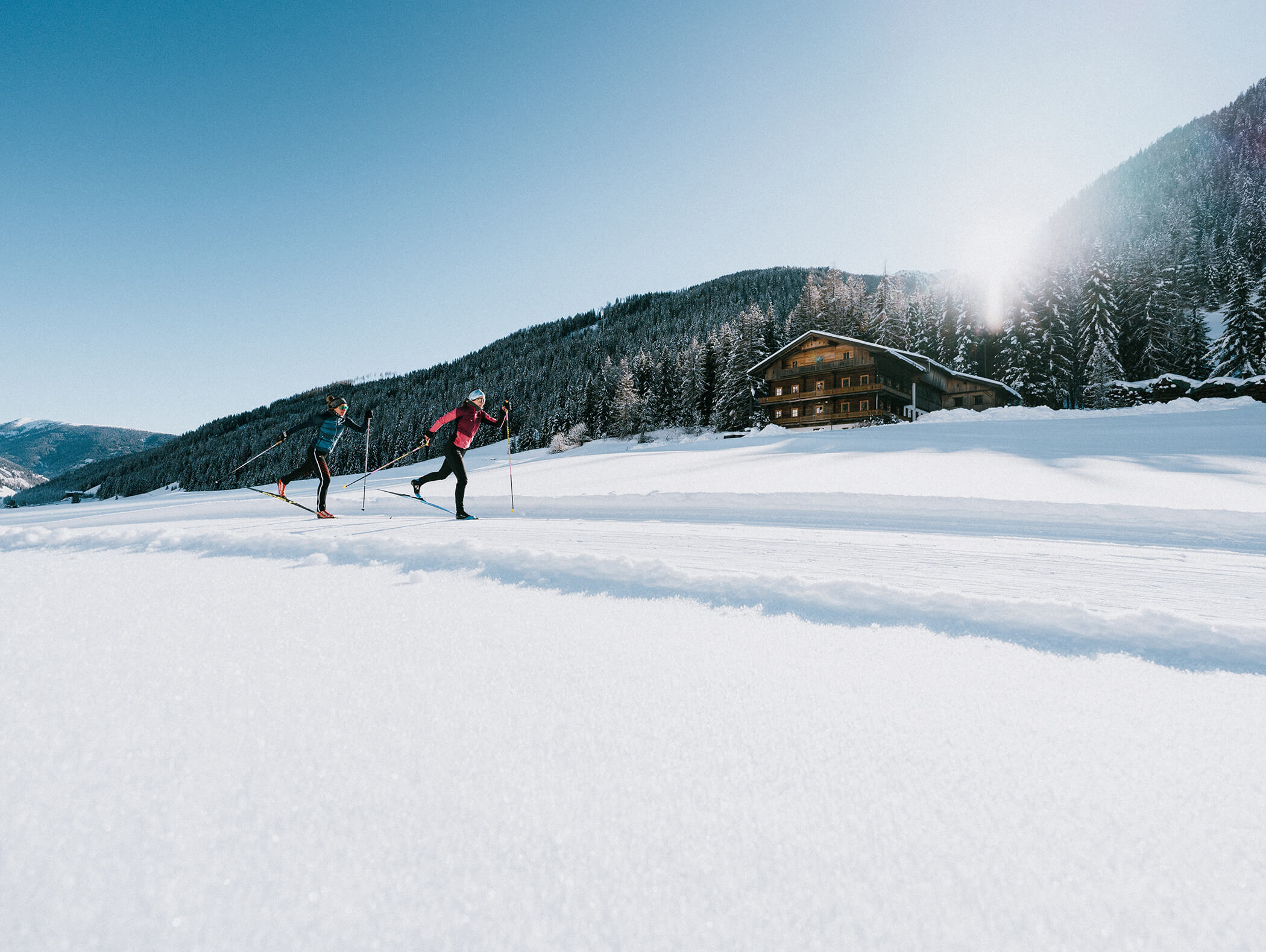 Zwei Langläufer auf Schnee in der Nähe einer Holzhütte, mit Bergen und Kiefern im Hintergrund. - AMIL chalets dolomites