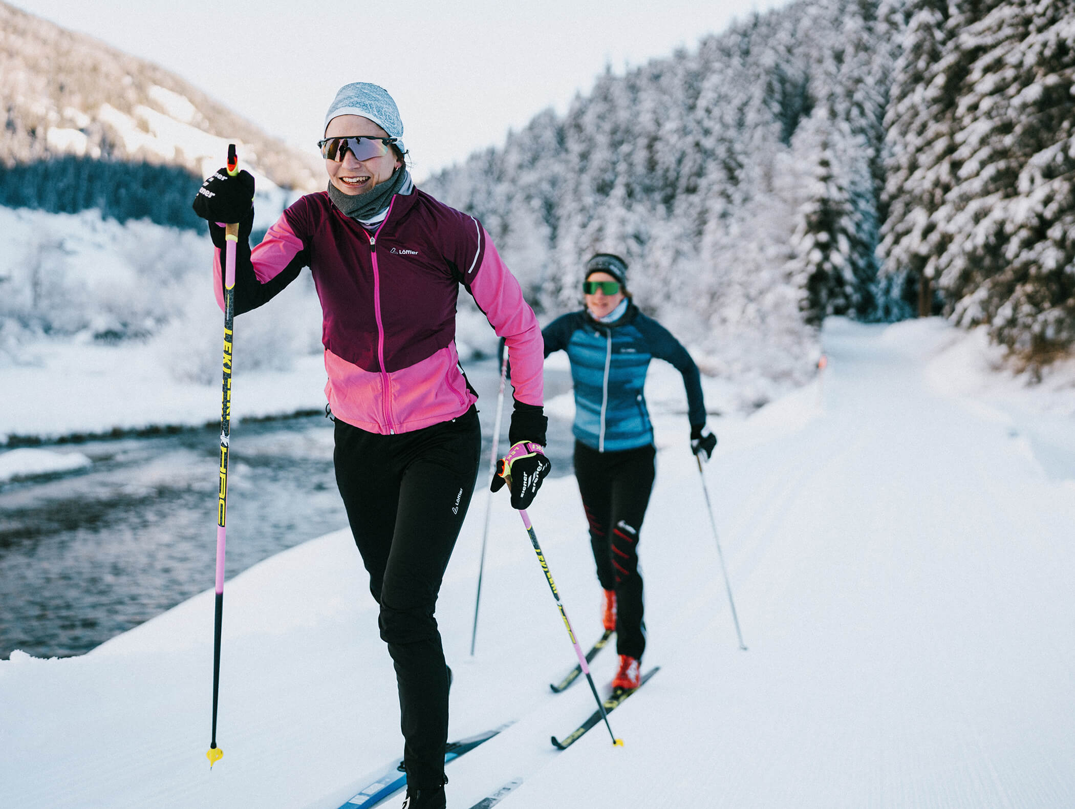 Zwei Personen fahren Langlauf auf einer verschneiten Loipe neben einem Fluss, umgeben von schneebedeckten Bäumen. - AMIL chalets dolomites