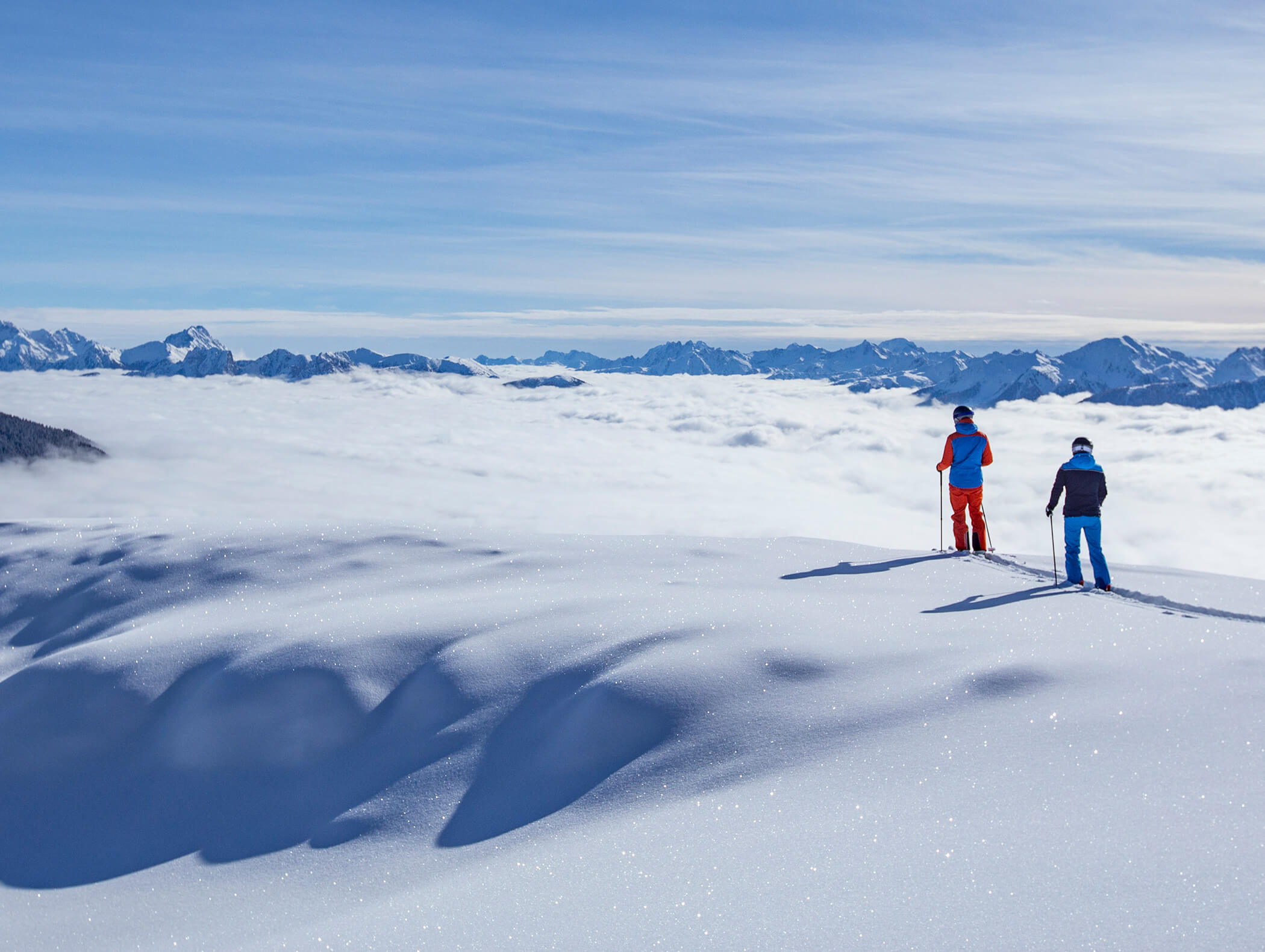 Zwei Skifahrer stehen an einem verschneiten Berghang und blicken auf ein Wolkenmeer und ferne Berggipfel. - AMIL chalets dolomites
