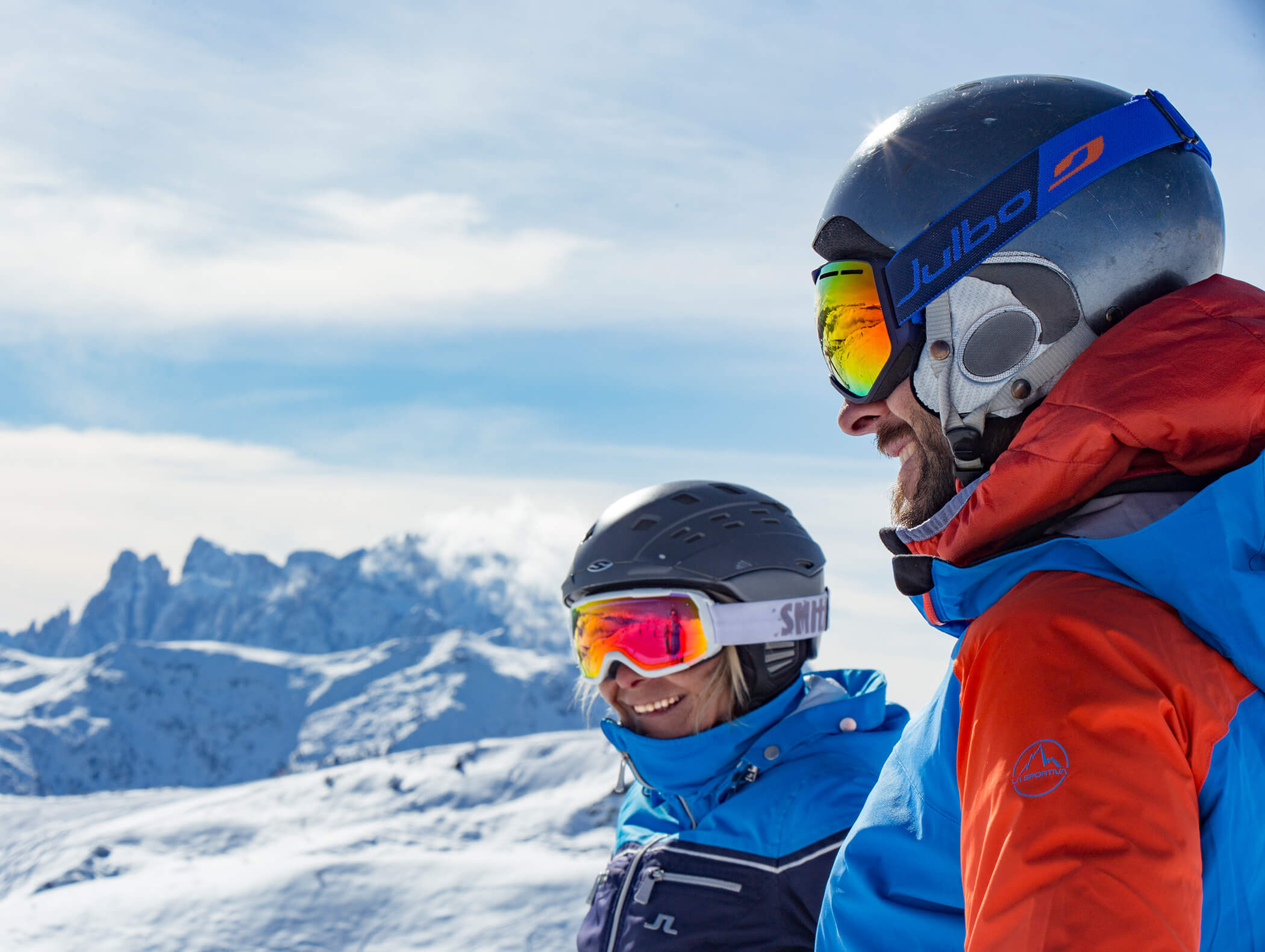 Zwei Skifahrer mit Helmen und Skibrillen lächeln auf einem verschneiten Berg mit Gipfeln im Hintergrund. - AMIL chalets dolomites