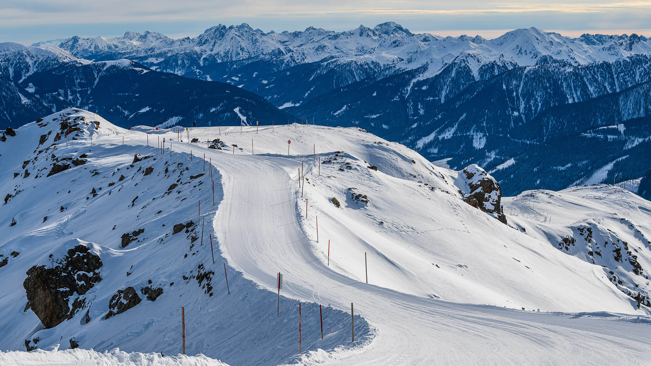 Verschneiter Berghang mit gewundener Skipiste vor der Kulisse der verschneiten Gipfel in der Ferne. - AMIL chalets dolomites