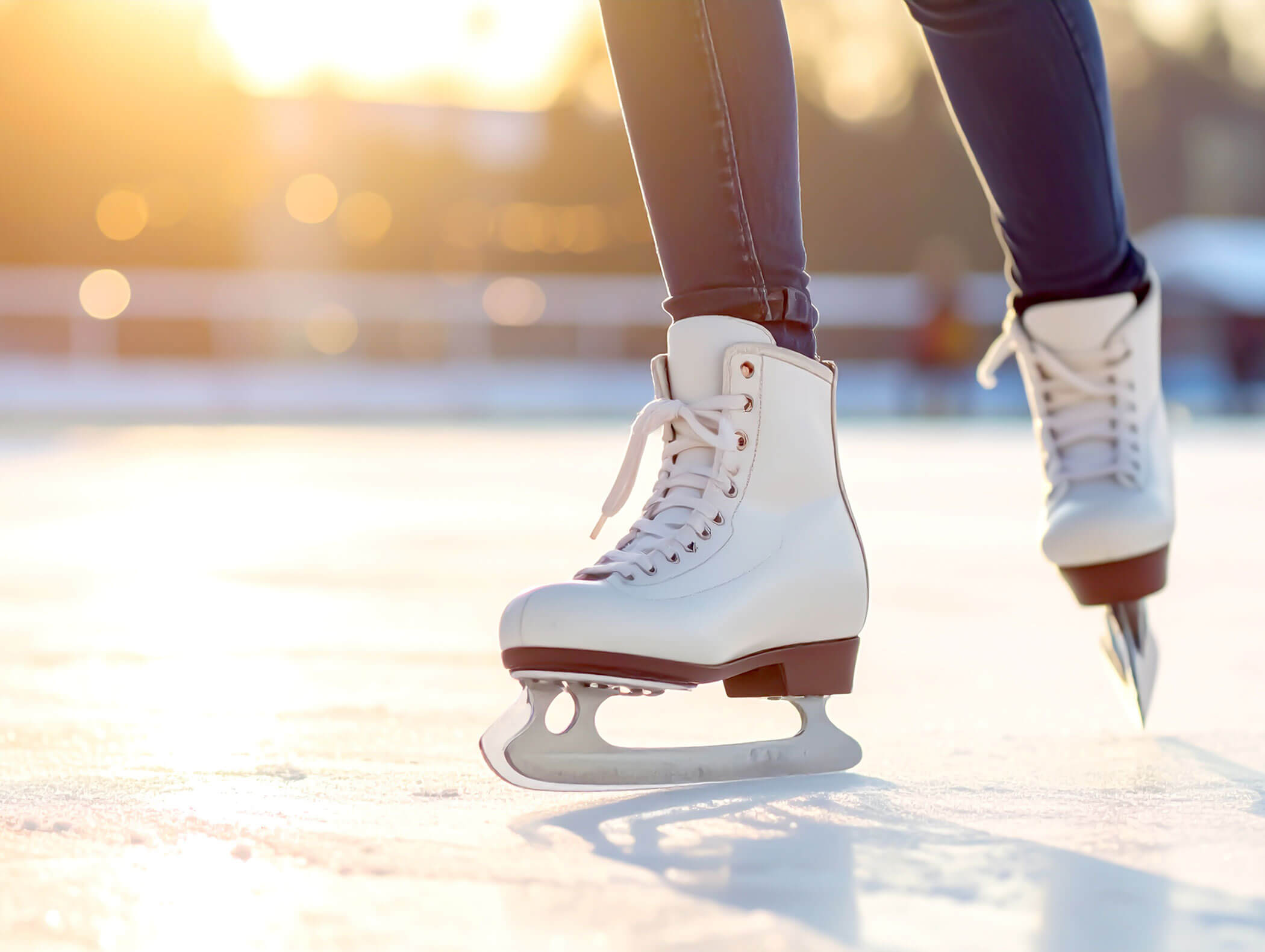Nahaufnahme einer Person mit weißen Schlittschuhen, die auf einer Eisbahn im Freien im Sonnenlicht gleitet. - AMIL chalets dolomites