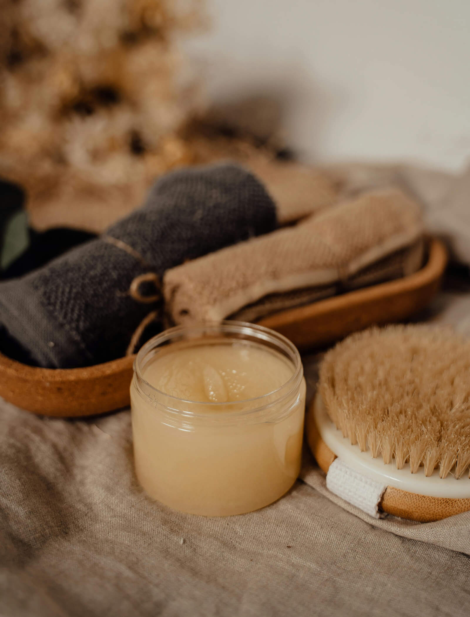 A jar of body scrub, a dry brush, and rolled towels arranged on a neutral fabric surface. - AMIL chalets dolomites