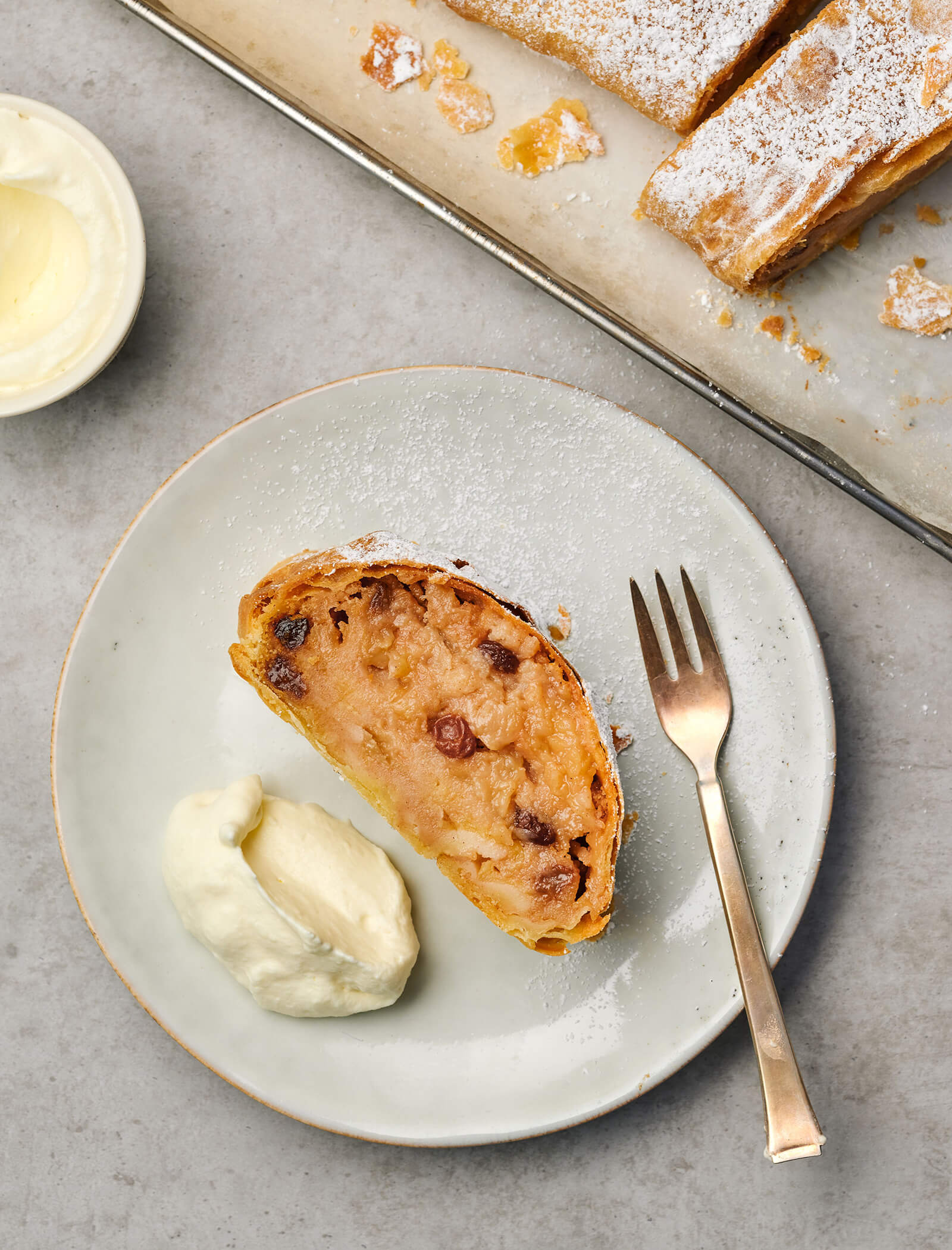 Una fetta di strudel di mele con uvetta su un piatto, servita con panna montata e una forchetta. - AMIL chalets dolomites