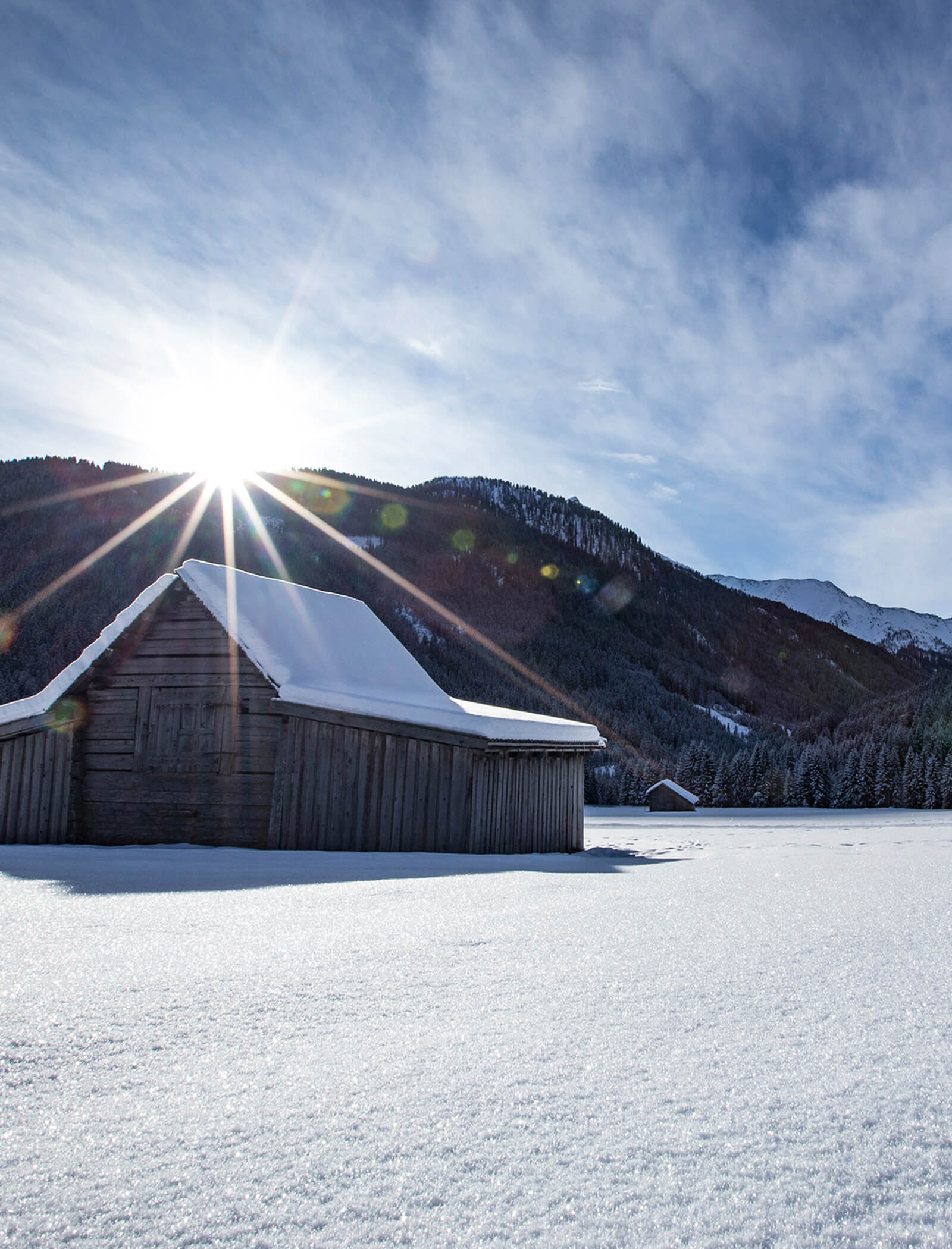 A wooden cabin with a snowy roof stands in a snow-covered field at sunrise, with mountains in the background. - AMIL chalets dolomites